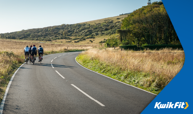 A group of cyclists on a countryside road surrounded by trees, fields and hills with a bright blue sky..