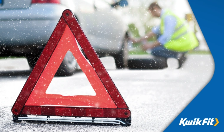 Red reflective warning triangle placed behind car in the snow while a wheel gets replaced in the distance.