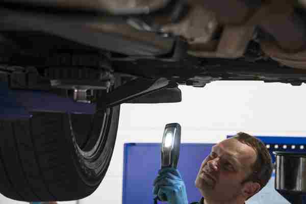 A man wearing blue gloves standing underneath a car looking at the steering and suspension with a torch 